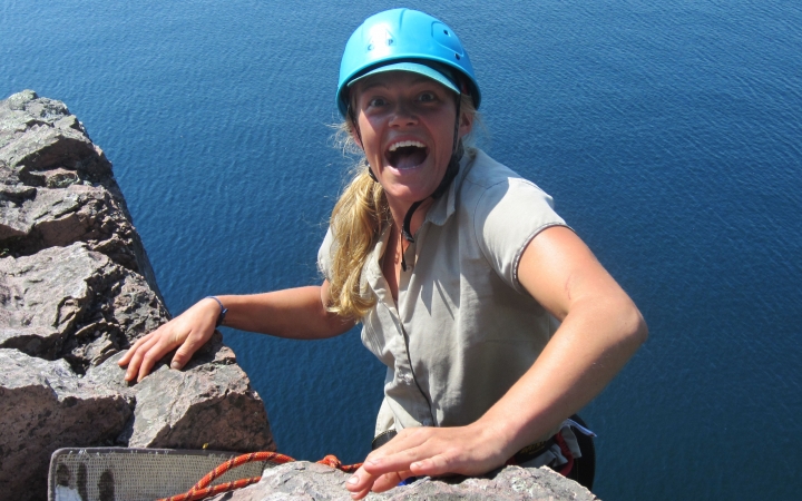 A person wearing safety gear is secured by ropes as they stand on the edge of a cliff high above a blue body of water and smile excitedly. 